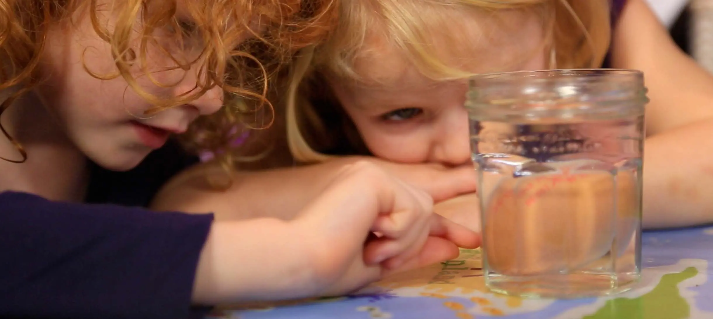 two young girls leaning over a table looking at an unpeeled egg floating in a glass jar