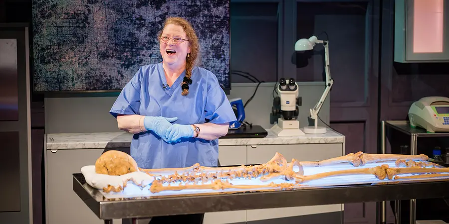 Sue Black is standing behind a table with a skeleton laid out on it, smiling. She is wearing blue scrubs and gloves.