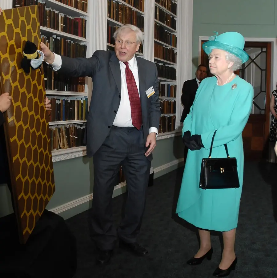 The queen wearing a green dress and hat with David Attenborough in a corridor looking at a painting of honeycombs with a toy bee