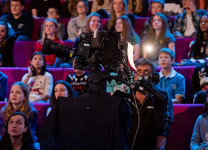 A TV camera is in the foreground with children and adults sitting in the background watching something we cannot see