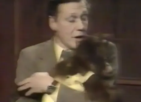 David Attenborough holding a monkey, who is wearing a yellow dungarees