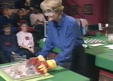Nancy Rothwell with chemistry goggles on, holding a set beakers on a green table