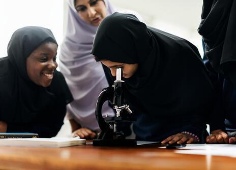 A group of hijabi girls smiles as one looks into a microscope