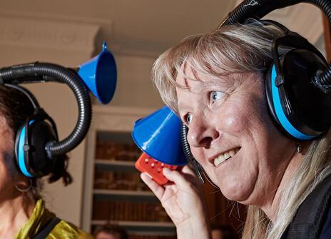 blond woman holding one end of a plastic trombone like device that is sitting on her head, she appears to be listening and smiling