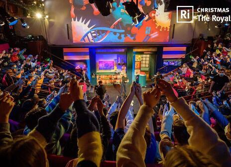 Young people with their hands up during the Christmas Lectures filming in the Ri Theatre