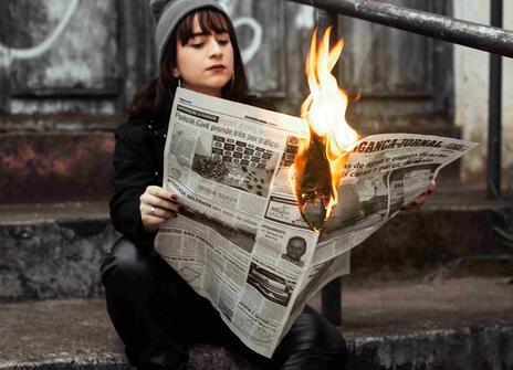 A girls reads a newspaper that is on fire in her hands